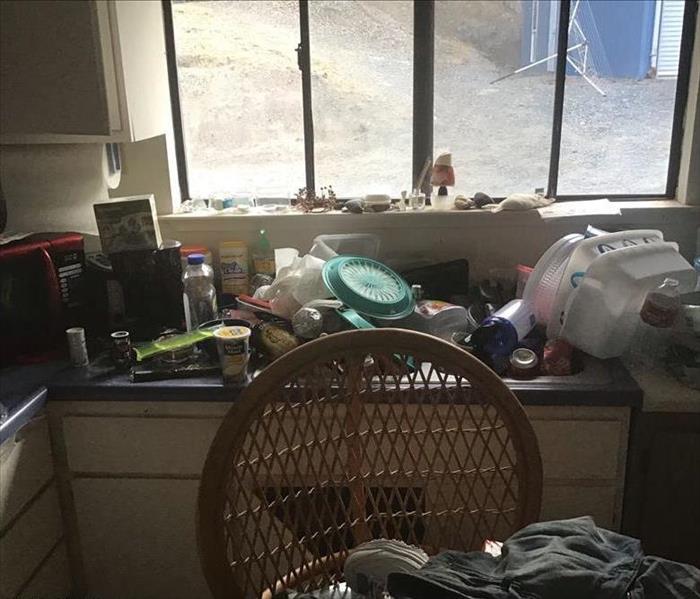 kitchen area filled with clutter from plates, empty containers, boxes. Clothes piled on chair